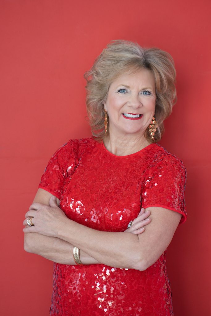 Portrait of author and speaker Marita Littauer smiling in a red dress with her arms folded against a red background.